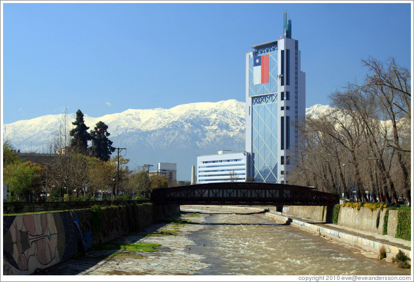 Looking east along the Mapocho River toward the mountains and a building with a Chilean flag.