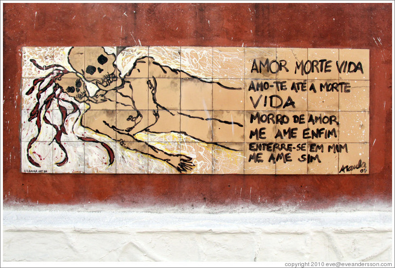 Artwork: lovers with skulls for heads.  The accompanying text reads: Amor Morte Vida / Amo-te At? Morte / Vida / Morro de Amor / Me Ame Enfim / Enterre-se em Mim / Me Ame Sim.  Wall surrounding the Cemit?o S?Paulo.