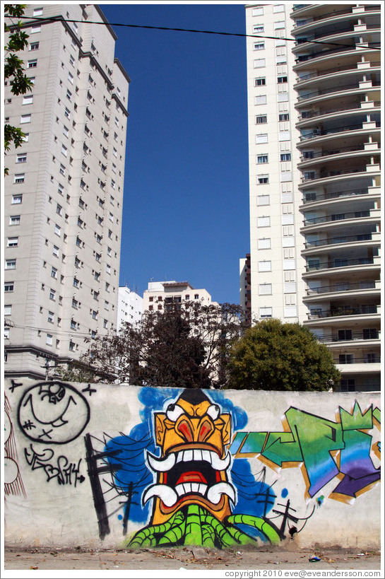 Graffiti: creature with pointed teeth and green tubes.  Buildings in the background.  Av. H?o Pellegrino at Rua Marcos Lopes.