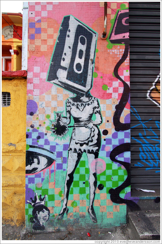 Graffiti: woman with a cassette tape head wearing a sexy maid outfit.  On her left, the head of a woman with a crown.  Villa Magdalenda neighborhood.  Rua Cardeal Arcoverde.