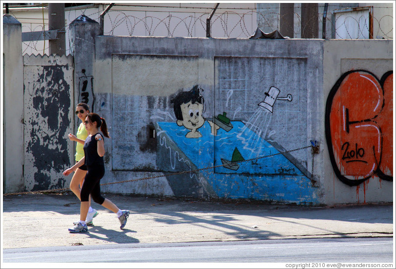 Graffiti: kid in a bathtub with boats. Two women walking past.  Rua Manuel da N?ga at Av. Mal. Est?o Albuquerque Lima.
