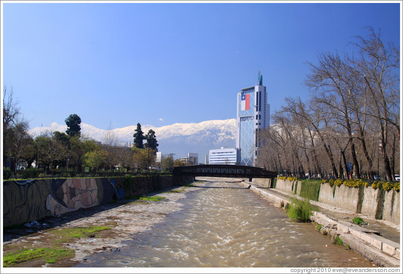 Looking east along the Mapocho River toward the mountains and a building with a Chilean flag.