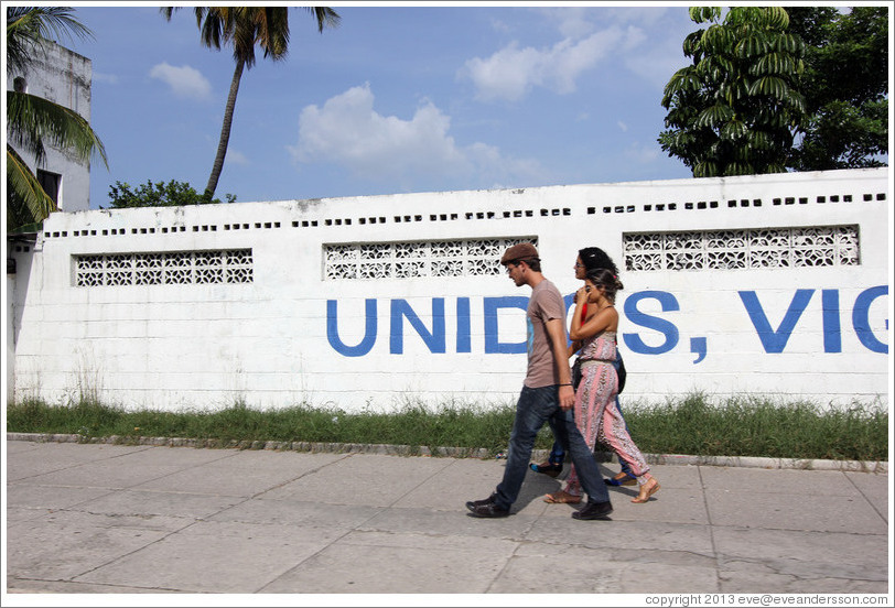 Couple walking past a wall painted with the word "Unidos", part of "Unidos, Vigilantes, y Combativos" ("United, Vigilant, and Combative").