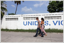 Couple walking past a wall painted with the word "Unidos", part of "Unidos, Vigilantes, y Combativos" ("United, Vigilant, and Combative").