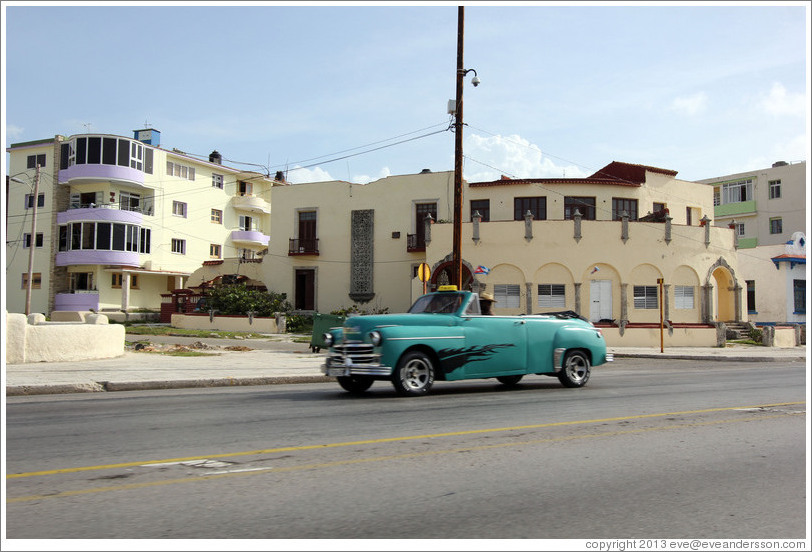 Green car passing beige buildings on the Malec&oacute;n.