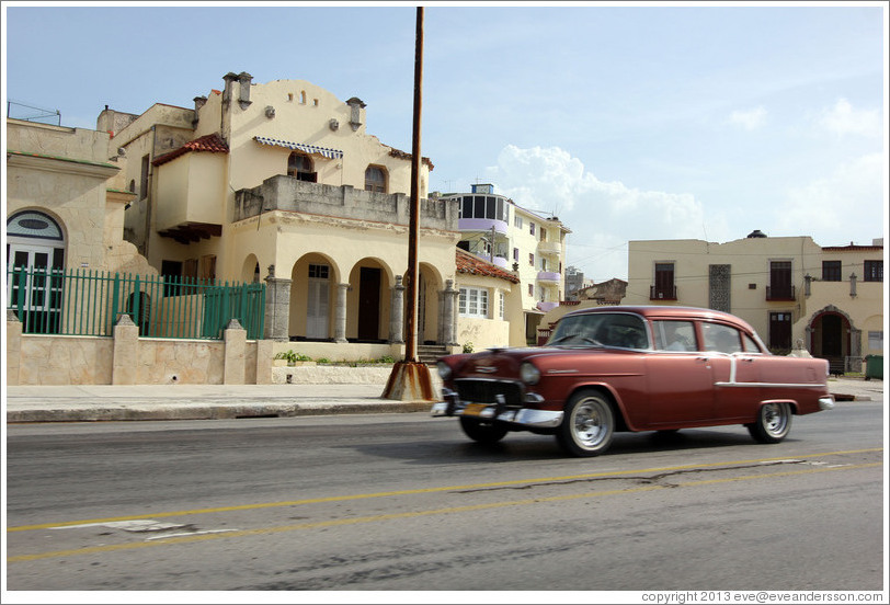 Red car passing beige buildings on the Malec&oacute;n.