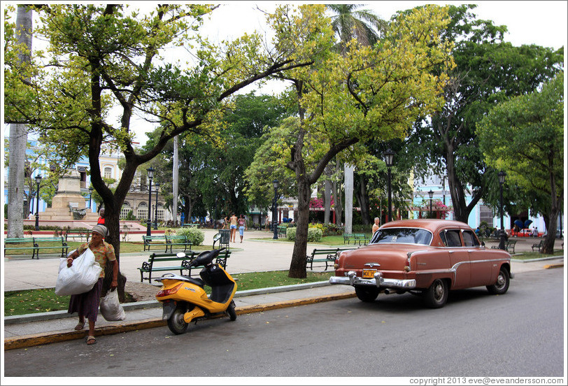 Woman carrying bags next to a yellow scooter and red car, Parque de la Libertad (Liberty Park).