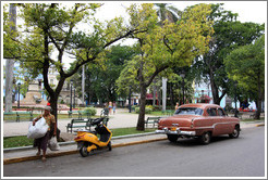 Woman carrying bags next to a yellow scooter and red car, Parque de la Libertad (Liberty Park).