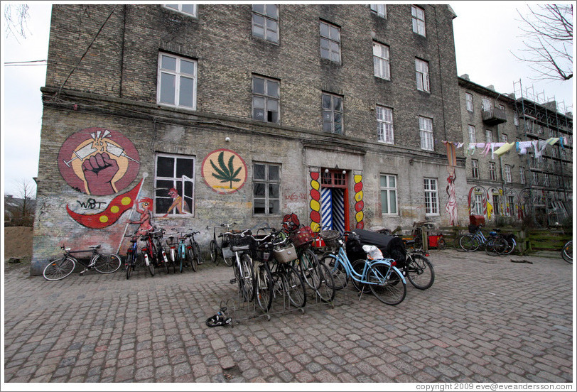 Paintings on a building, including a needle being broken, a woman with a Christiania flag, and a marijuana leaf.