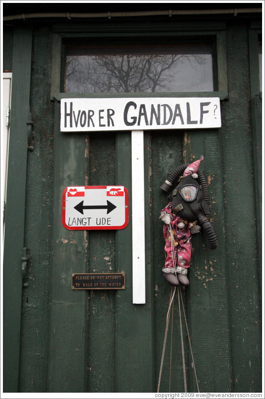 Door with sign saying "Hvor er Gandalf?" ("Where is Gandalf?"), another saying "langt ude" ("long way"), a doll in a gas mask, and a 3rd sign saying "Please do not attempt to walk on the water".