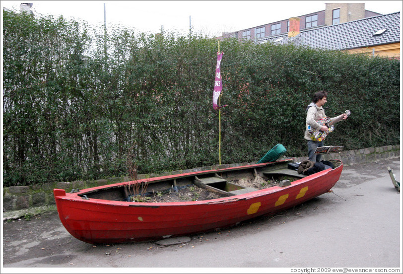 Guitar player, near a wooden boat painted with the three dots of Christiania.