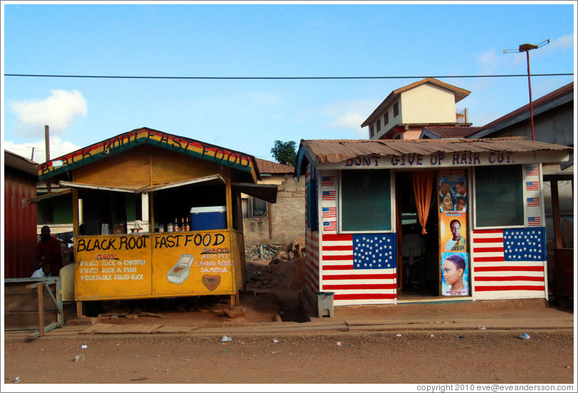 Two roadside shops Black Root Fast Food and Don't Give Up Hair Cut.  The latter is plastered with American flags.