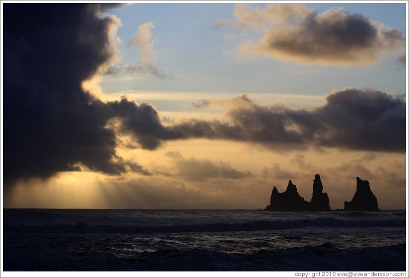Reynisdrangar, volcanic rock shooting from the ocean, with the setting sun shining from behind clouds.