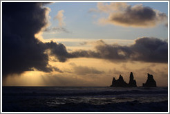 Reynisdrangar, volcanic rock shooting from the ocean, with the setting sun shining from behind clouds.