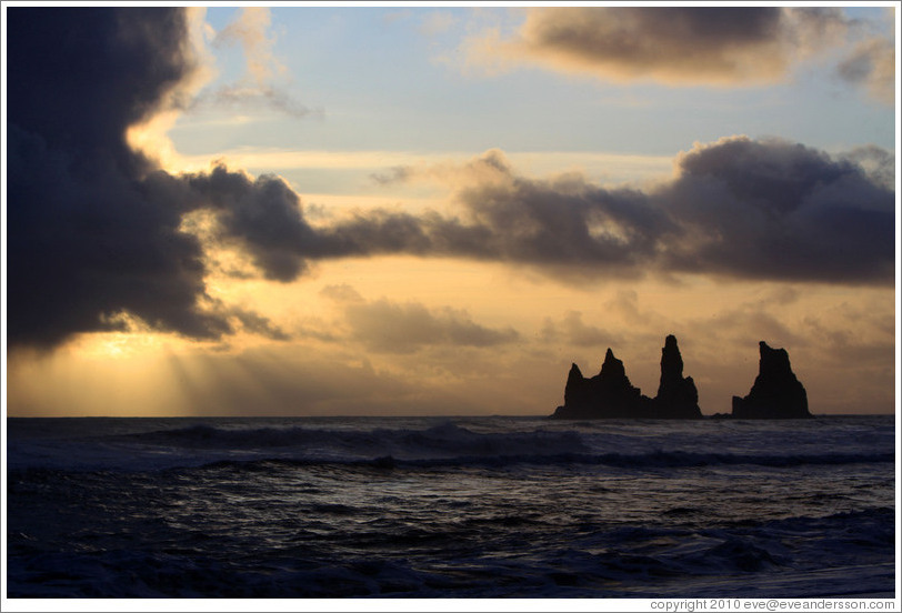 Reynisdrangar, volcanic rock shooting from the ocean, with the setting sun shining from behind clouds.