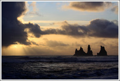 Reynisdrangar, volcanic rock shooting from the ocean, with the setting sun shining from behind clouds.