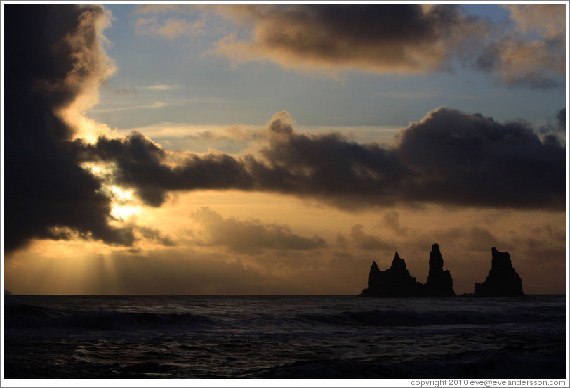 Reynisdrangar, volcanic rock shooting from the ocean, with the setting sun shining from behind clouds.