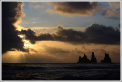 Reynisdrangar, volcanic rock shooting from the ocean, with the setting sun shining from behind clouds.