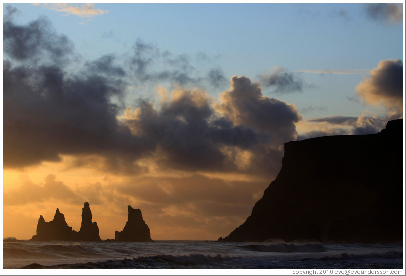 Reynisdrangar, volcanic rock shooting from the ocean, under the mountain Reynisfjall, with the setting sun shining from behind clouds. 