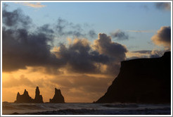 Reynisdrangar, volcanic rock shooting from the ocean, under the mountain Reynisfjall, with the setting sun shining from behind clouds. 