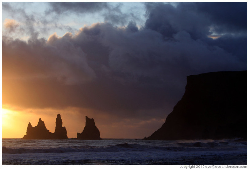 Reynisdrangar, volcanic rock shooting from the ocean, under the mountain Reynisfjall, with the setting sun shining from behind clouds.