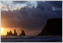 Reynisdrangar, volcanic rock shooting from the ocean, under the mountain Reynisfjall, with the setting sun shining from behind clouds.