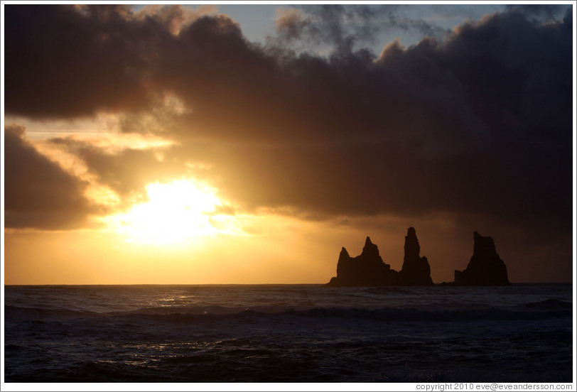 Reynisdrangar, volcanic rock shooting from the ocean, with the setting sun shining from behind clouds.