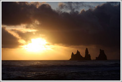 Reynisdrangar, volcanic rock shooting from the ocean, with the setting sun shining from behind clouds.