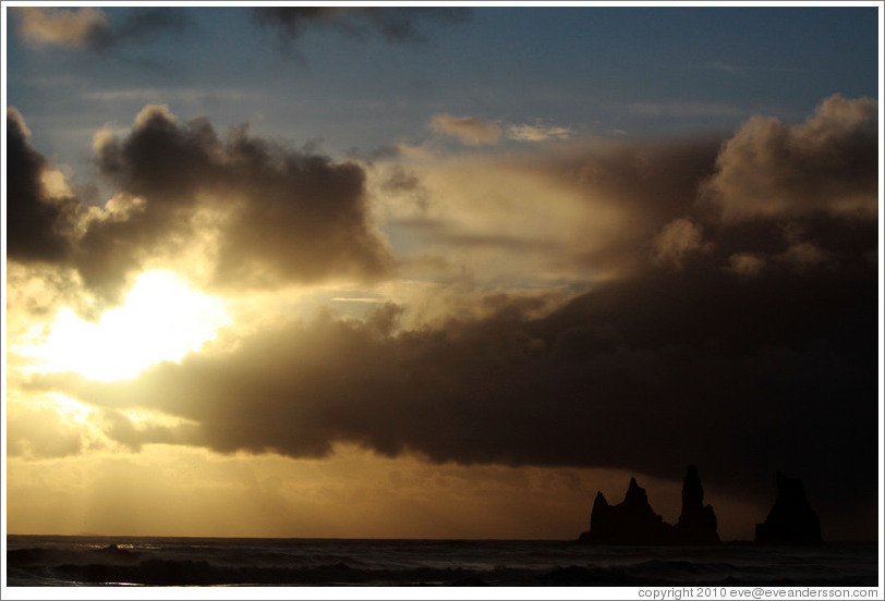 Reynisdrangar, volcanic rock shooting from the ocean, with the setting sun shining from behind clouds.