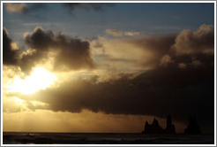 Reynisdrangar, volcanic rock shooting from the ocean, with the setting sun shining from behind clouds.