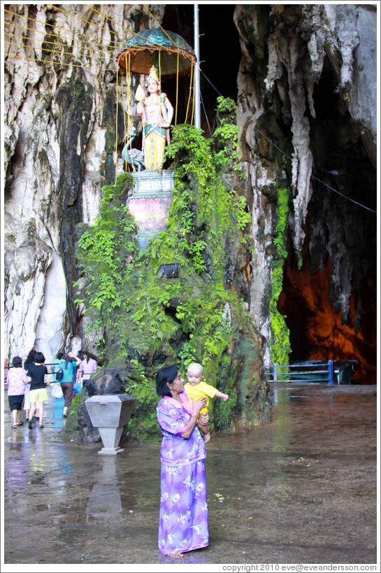 Woman and baby in front of statue of woman and peacock, Batu Caves.