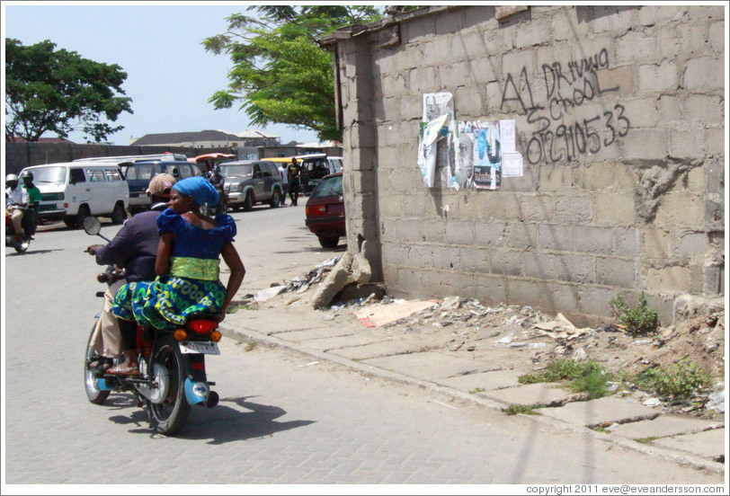 Couple on a motorcycle driving past A1 Driving School written on a wall. Victoria Island.
