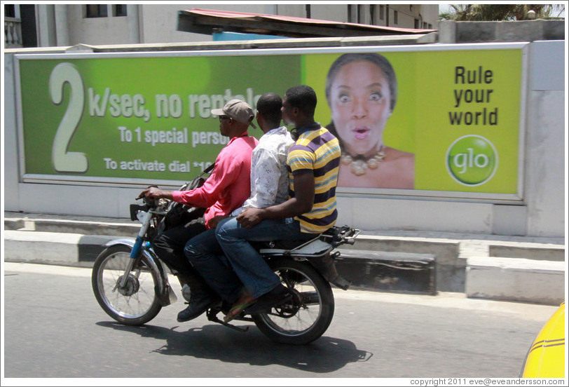 Three men on a motorcycle passing a glo billboard. Maroko Road, Victoria Island.