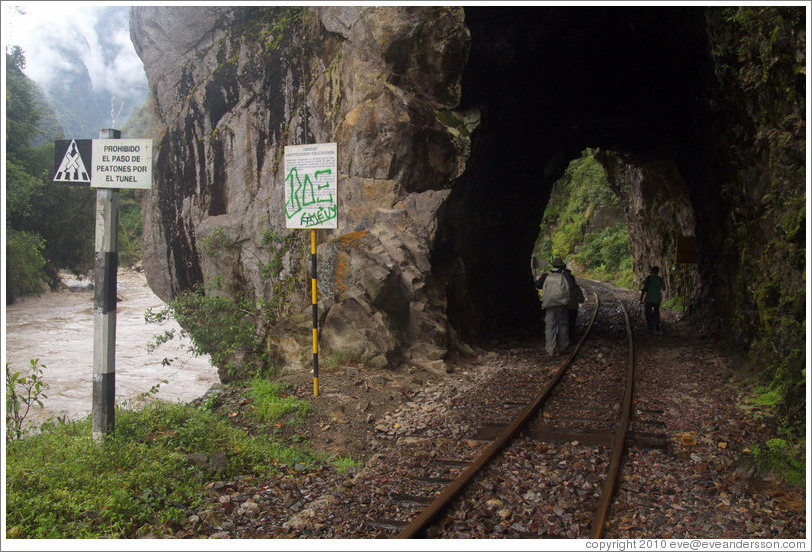 Pedestrains walking through a tunnel over the train tracks.  The sign reads "Prohibido el paso de patones por el tunel" ("The passage of pedestrians through the tunnel is prohibited.")
