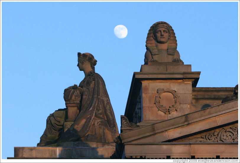 The moon appearing to hover near statues of a woman and a sphinx atop the Royal Scottish Academy&#8206; building.