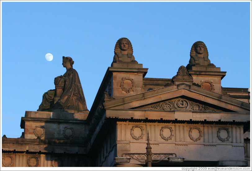 The moon appearing to hover near statues of a woman and sphinxes atop the Royal Scottish Academy&#8206; building.