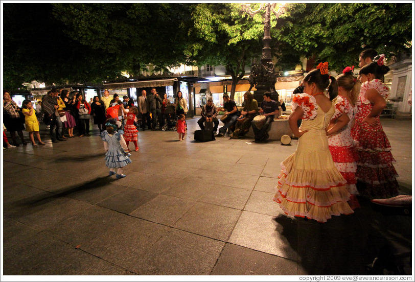 Three girls looking on street performers play music and others dance.  Fiesta de las Cruces.  Plaza de Bib-Rambla.  City center.
